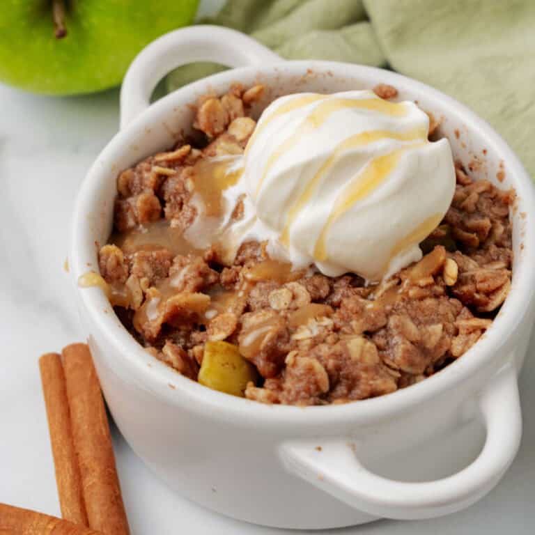 An apple crisp in a mug topped with whipped cream and caramel drizzle, with two cinnamon sticks and a green apple in the background.