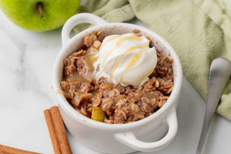 A small white bowl of apple crisp topped with a scoop of vanilla ice cream and caramel drizzle, next to a green apple, cinnamon sticks, and a metal spoon.