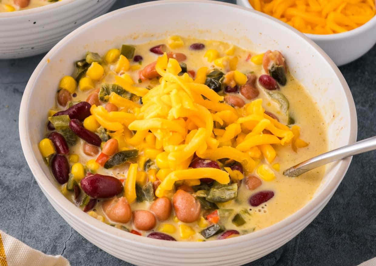 A bowl of creamy soup with beans, corn, diced vegetables, and a topping of shredded cheddar cheese. A spoon rests in the bowl.