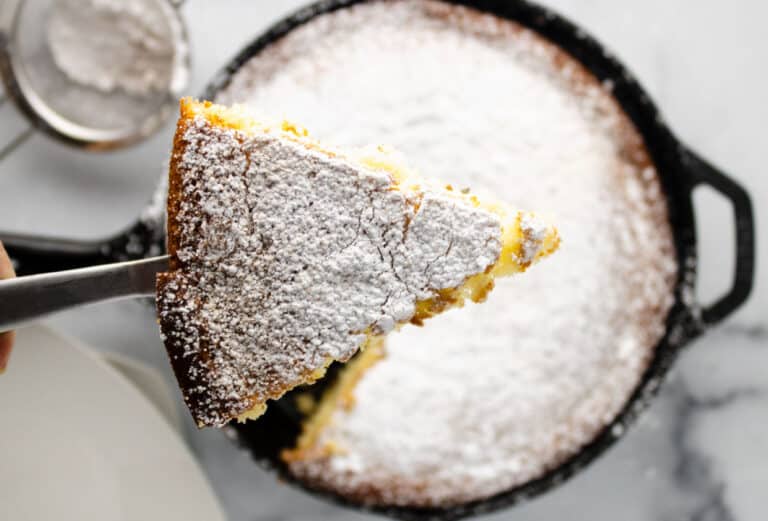 A close-up of a powdered sugar-dusted slice of cake being lifted from a cast iron skillet with a spatula.