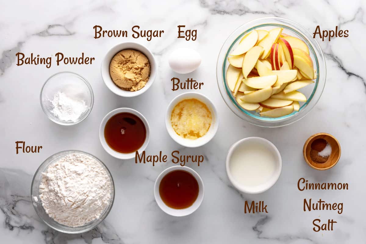 Top-down view of baking ingredients in bowls for an apple cinnamon cake, including flour, sugar, apple slices, butter, egg, milk, baking powder, maple syrup, cinnamon, nutmeg, and salt on a marble surface.