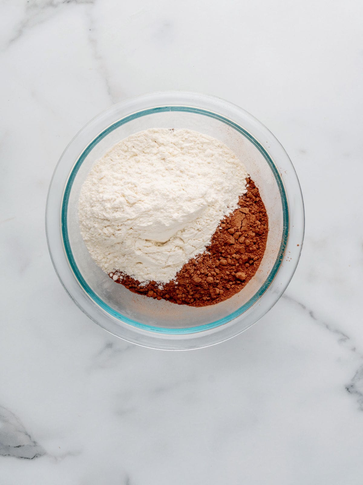 A clear glass bowl containing separate piles of flour and cocoa powder on a white marble surface.