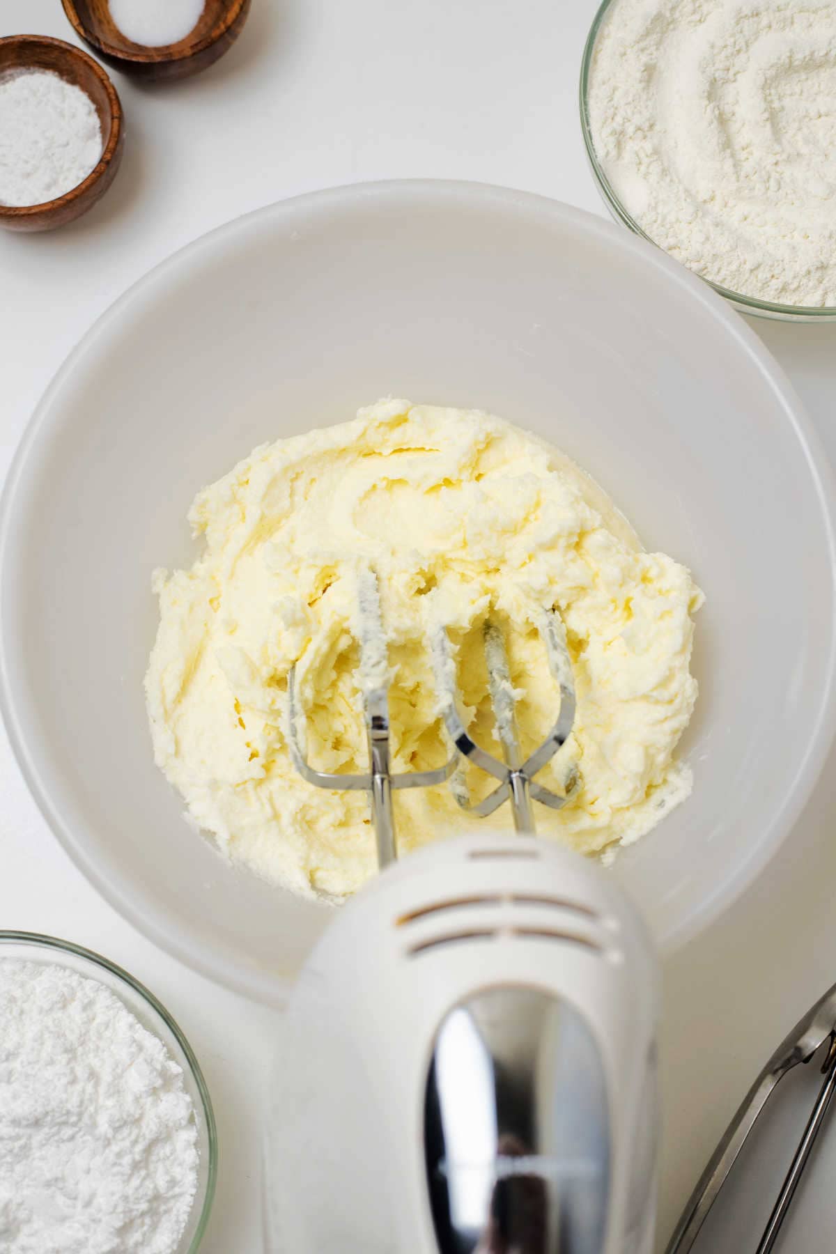 Electric hand mixer blending a creamy mixture butter and milk, surrounded by bowls of flour, powdered sugar, and salt on a white surface.