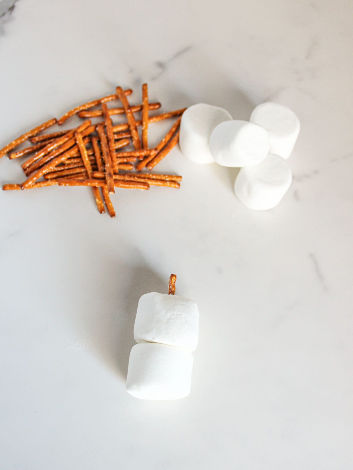 Two marshmallows stacked with a pretzel stick between them, with more pretzel sticks and marshmallows nearby on a white surface.