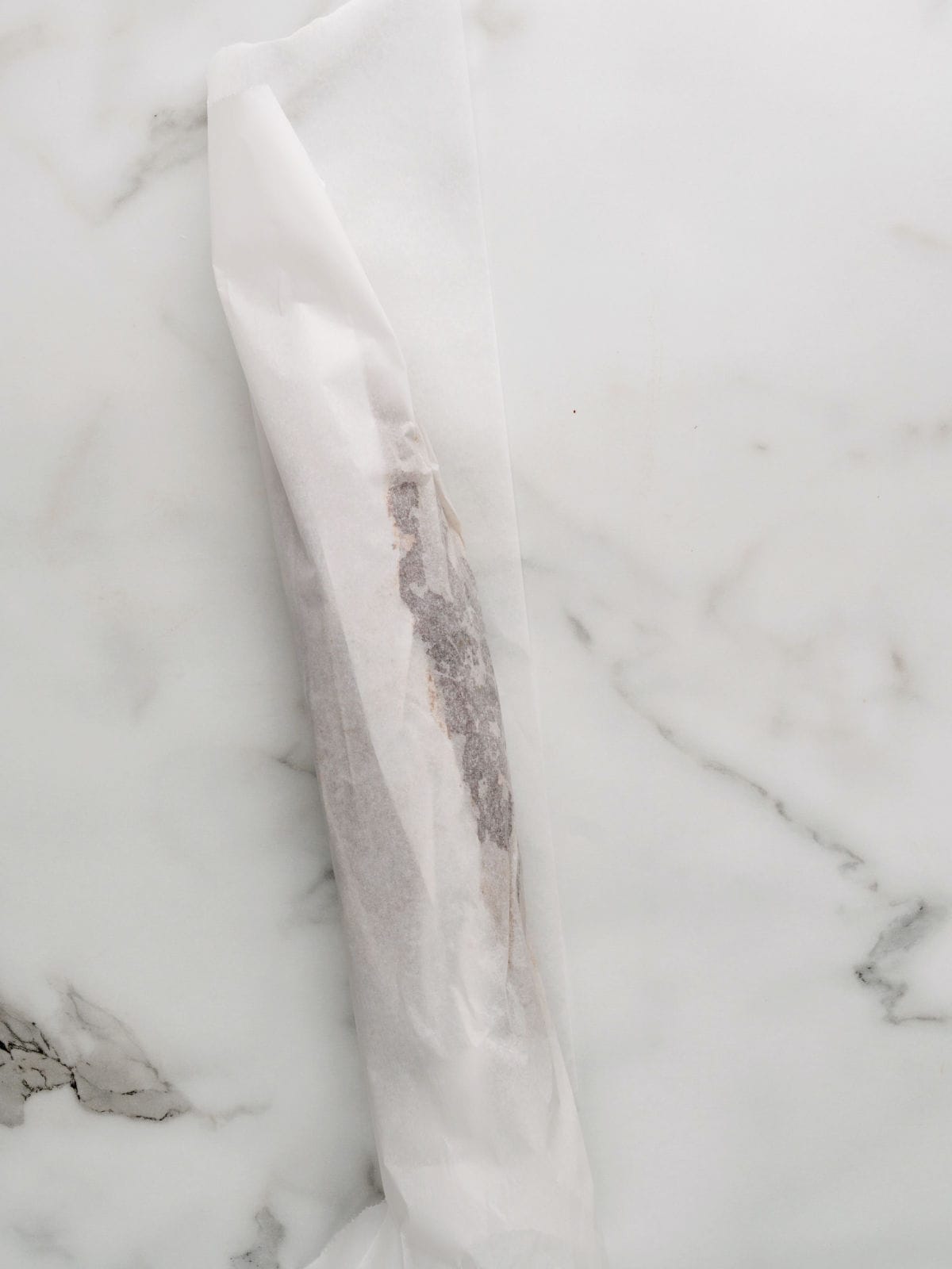 A long, thin log of chocolate cookie dough wrapped in white parchment paper lies on a white marble surface.