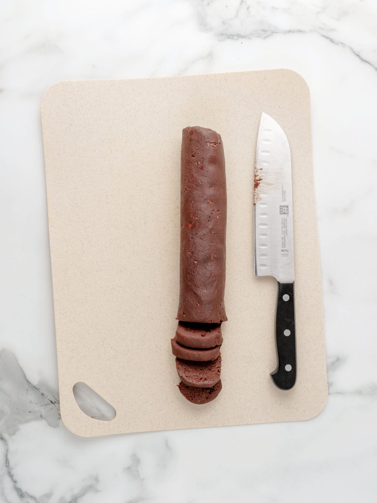 A log of chocolate cookie dough with cacoa nibs , slice and bake cookies is partially sliced on a beige cutting board next to a knife, all set on a marble countertop.