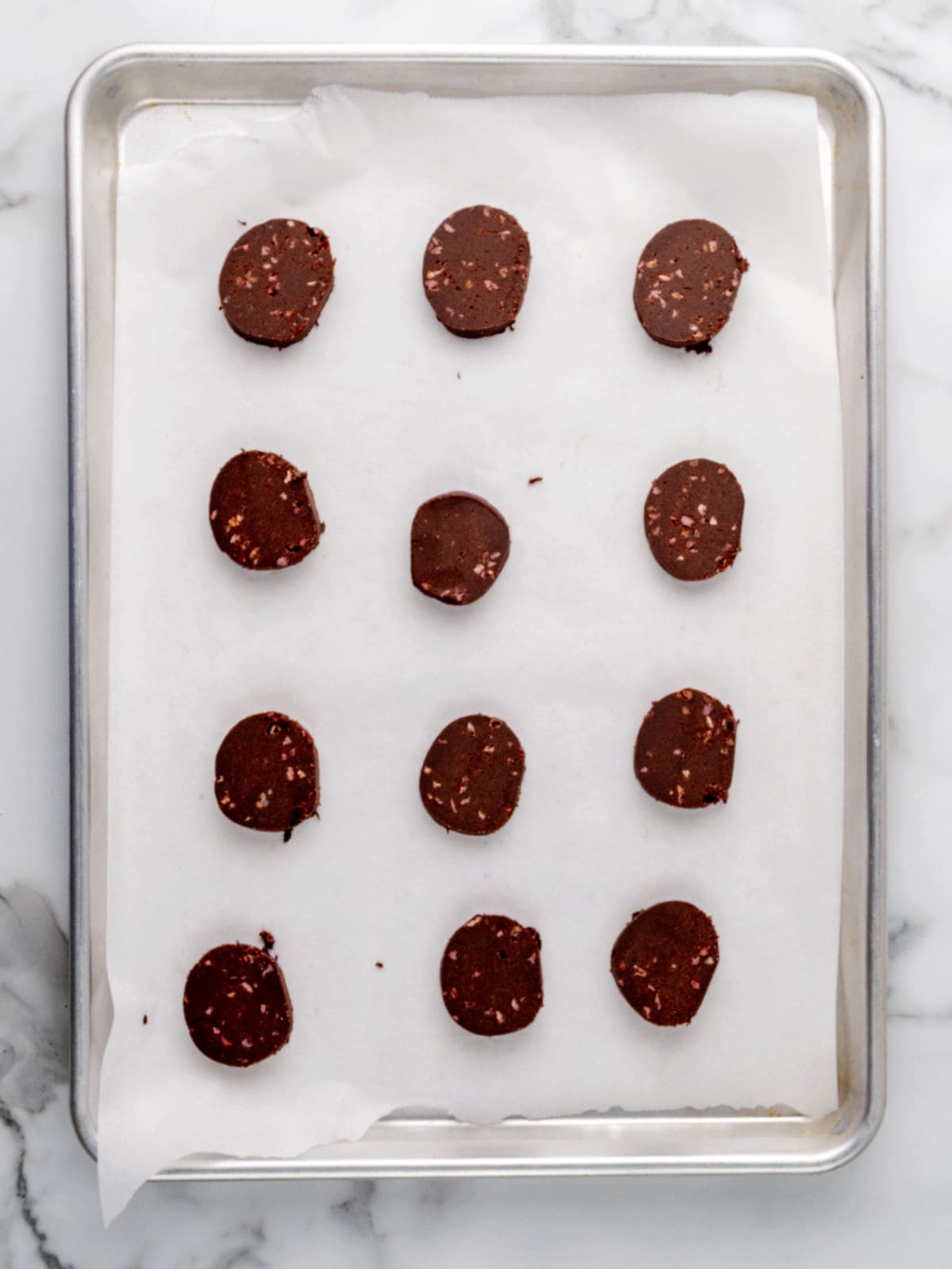 Twelve round slices of chocolate cookie dough are arranged on a parchment-lined baking sheet, ready to be baked.