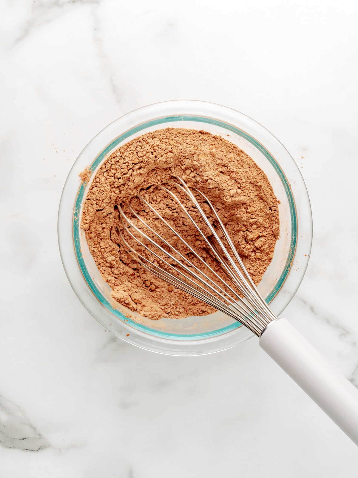A metal whisk rests in a glass bowl filled with cocoa powder mixture on a white marble surface.
