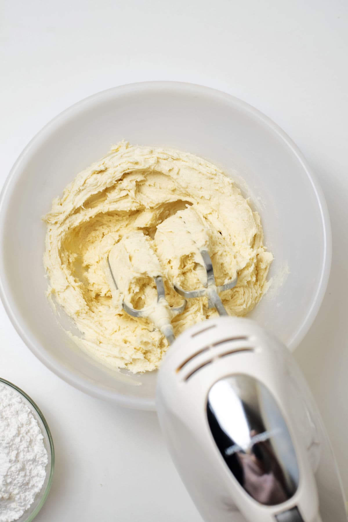 A hand mixer blending creamy batter for snowall cookies in a white bowl, with a small bowl of powdered sugar nearby on a white surface.