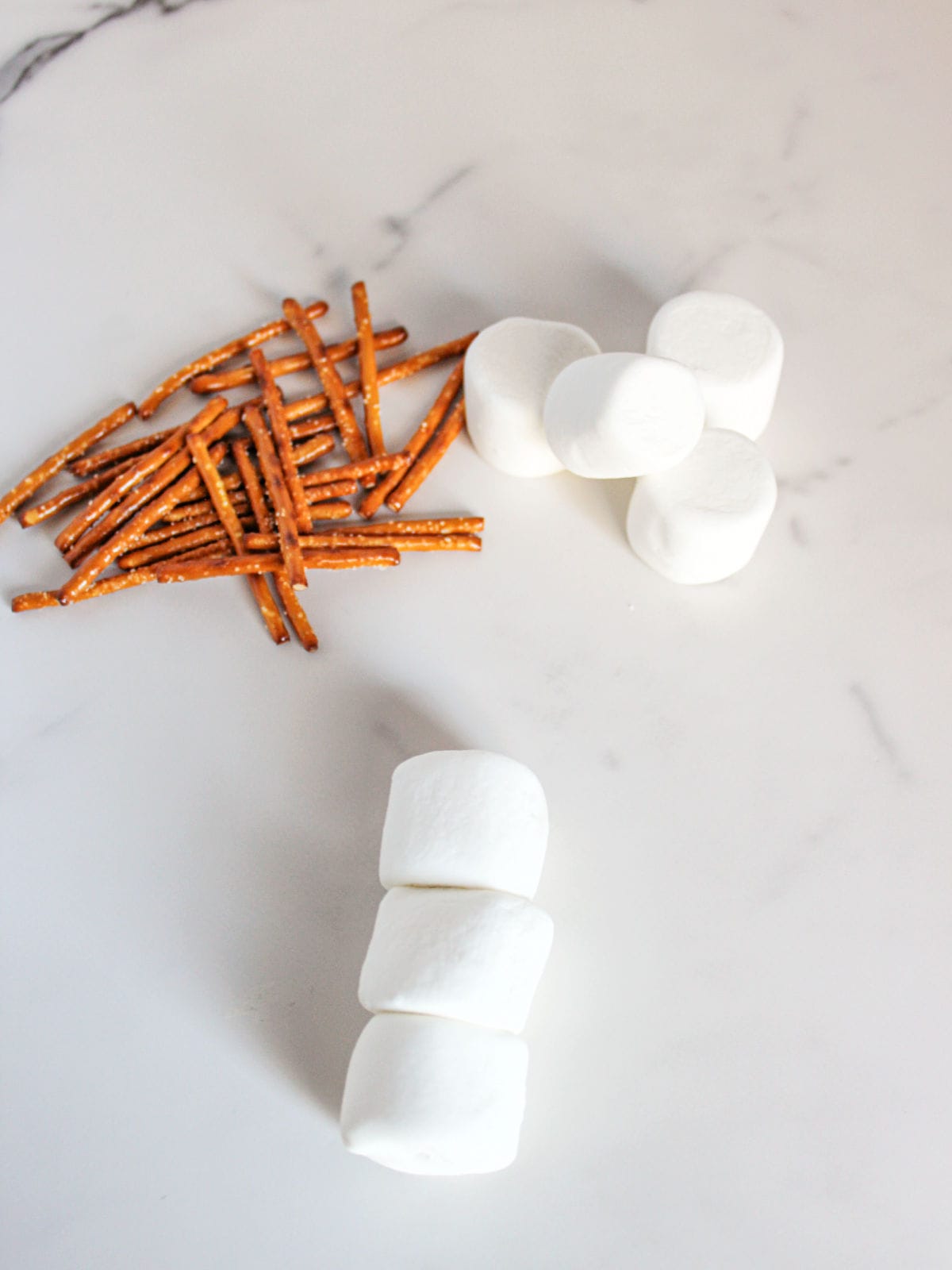 Three marshmallows stacked, four marshmallows in a cluster, and a pile of pretzel sticks on a white marble surface.