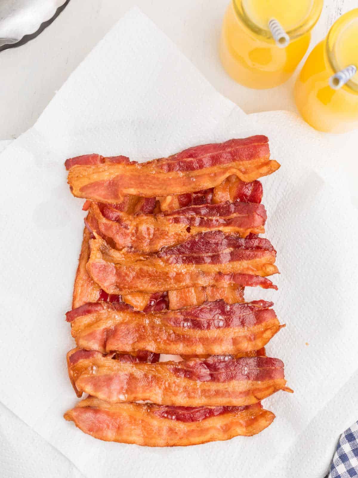 Eight cooked bacon strips rest on a paper towel, with two glass bottles of orange juice topped with striped straws nearby.
