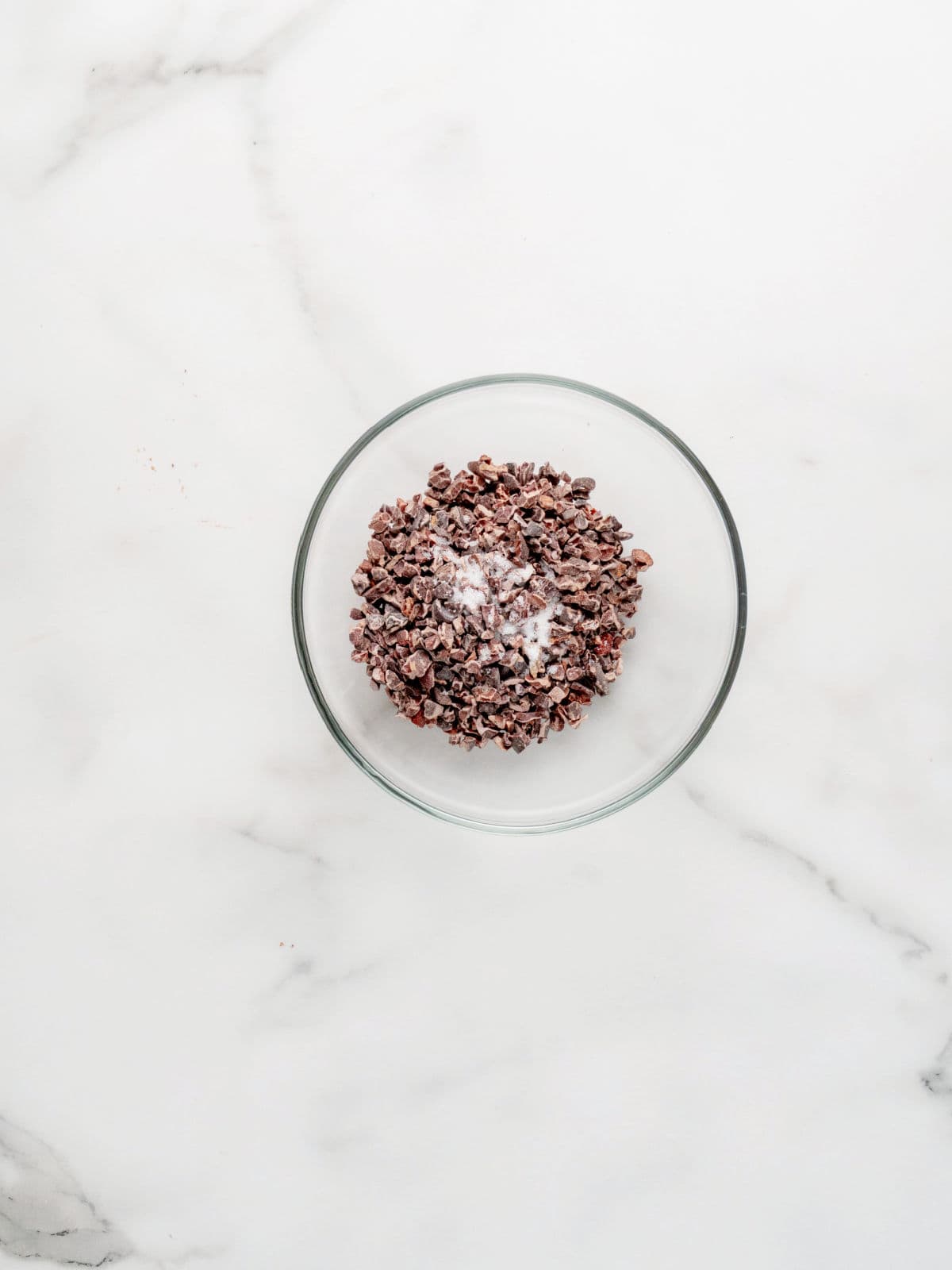 A glass bowl filled with cacao nibs and a small amount of white powder sits on a white marble surface.