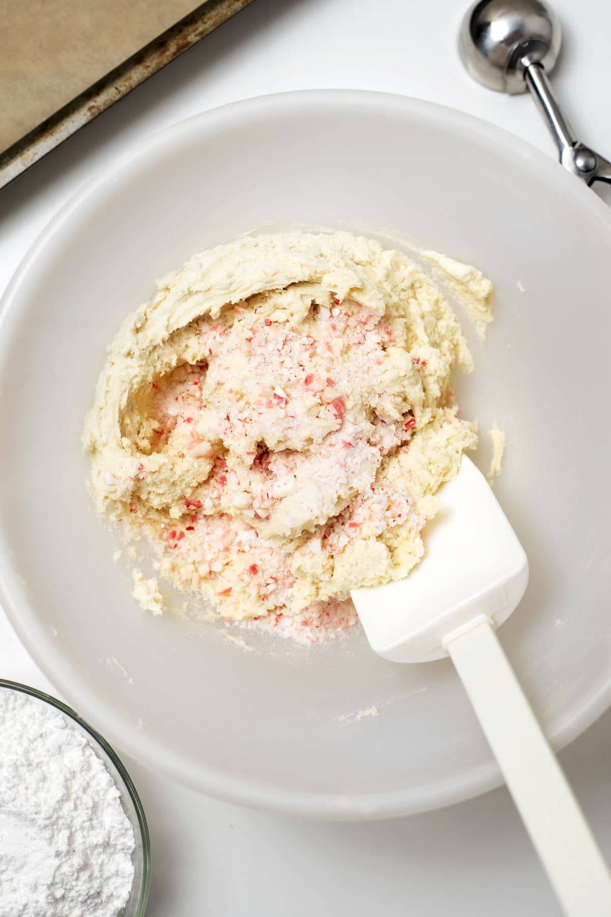 A mixing bowl with cookie dough and crushed peppermint being stirred with a white spatula; a scoop and a small bowl of powdered sugar are nearby.
