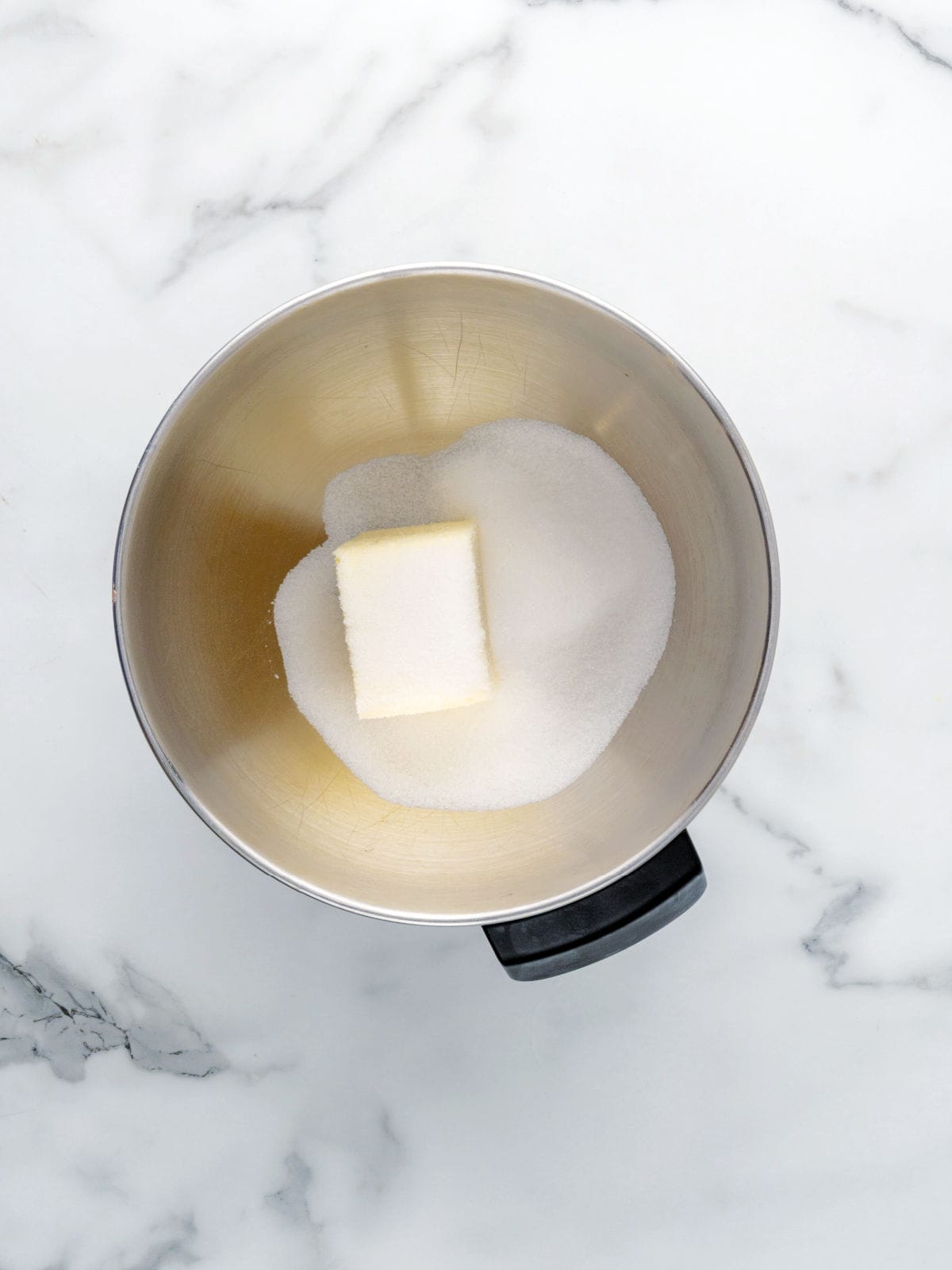 A metal mixing bowl containing a stick of butter and a pile of granulated sugar on a white marble surface.