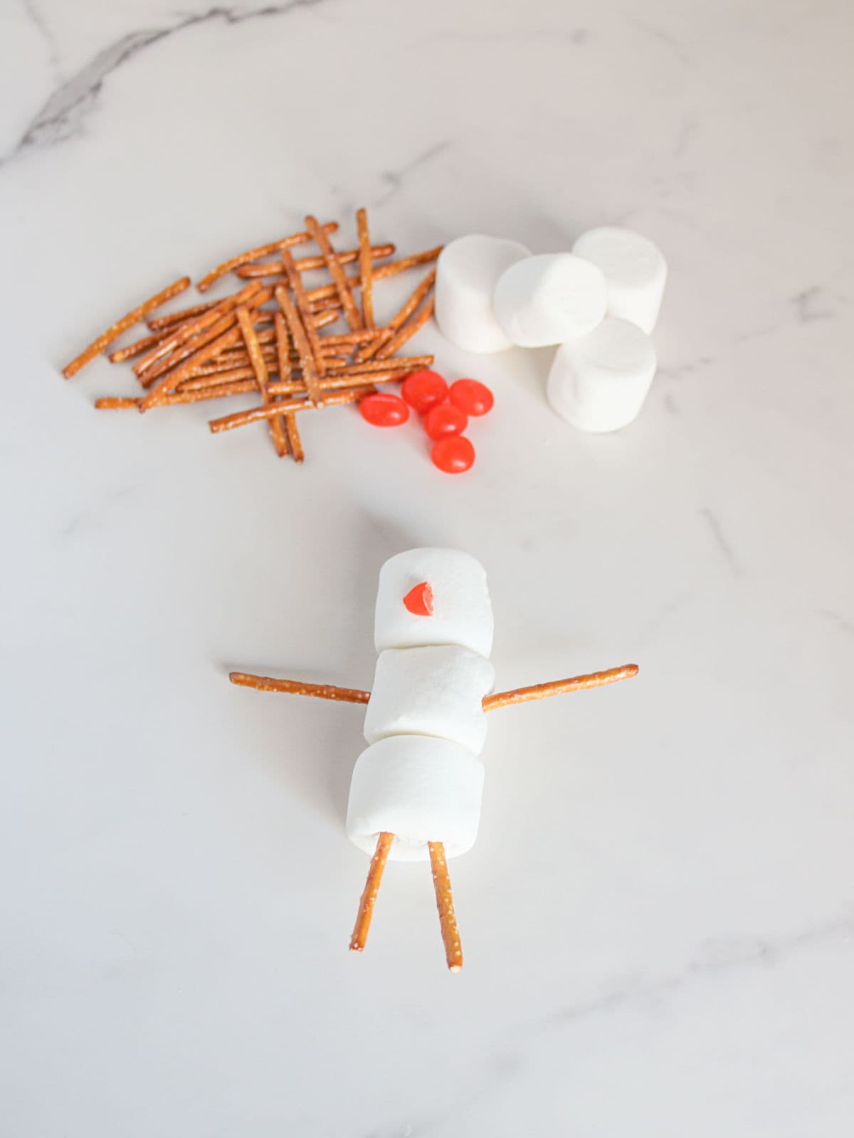 Marshmallows, pretzel sticks, and red candies are arranged on a white surface, with some pieces assembled to resemble a snowman.