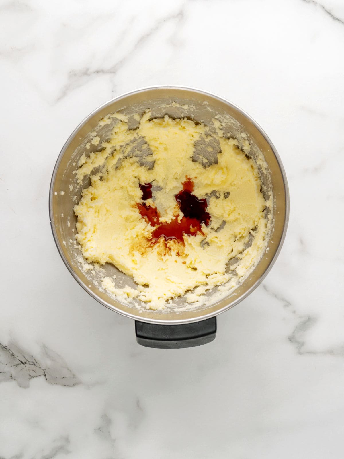 A metal mixing bowl with creamed butter and sugar, with a small amount of vanilla extract added on top, sits on a white marble surface.