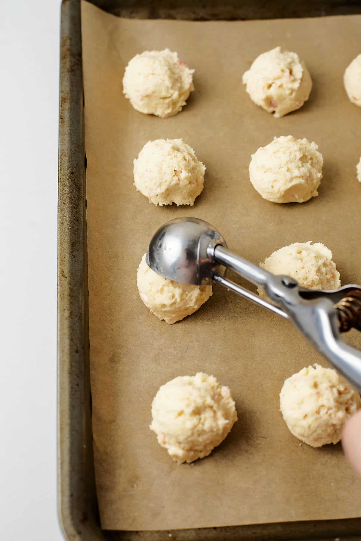 A hand uses a metal scoop to place balls of cookie dough onto a parchment-lined baking sheet, preparing them for baking.