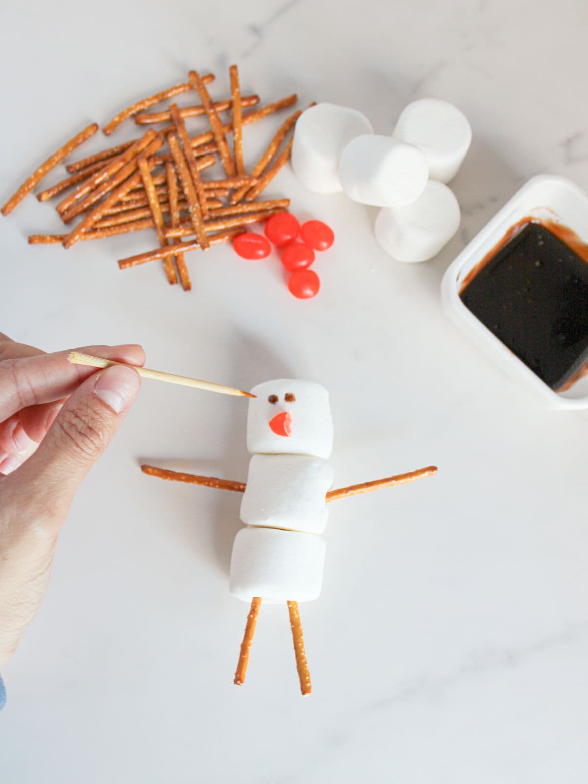 A hand decorates a snowman made of marshmallows and pretzel sticks, with red candy, chocolate sauce, and extra pretzels visible on a white surface.