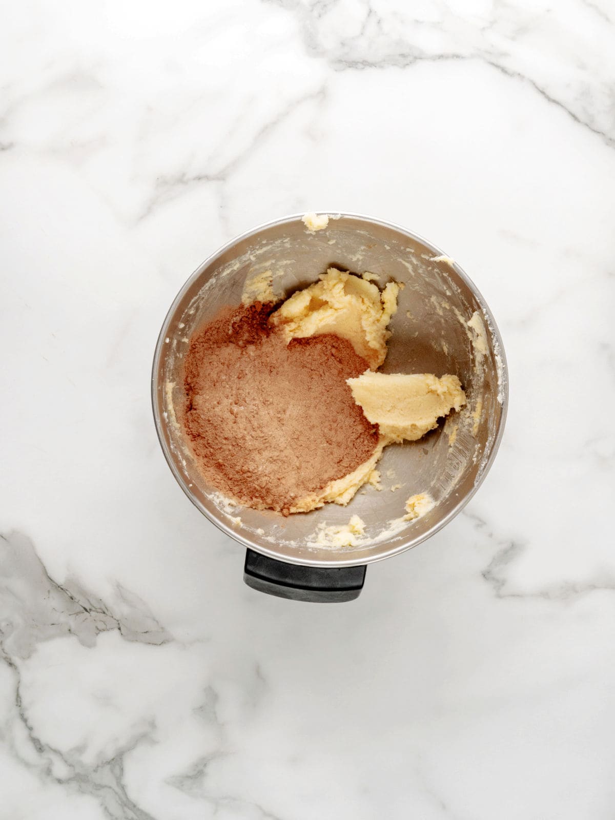 A metal mixing bowl with cookie dough and cocoa powder on a marble countertop, partially mixed.