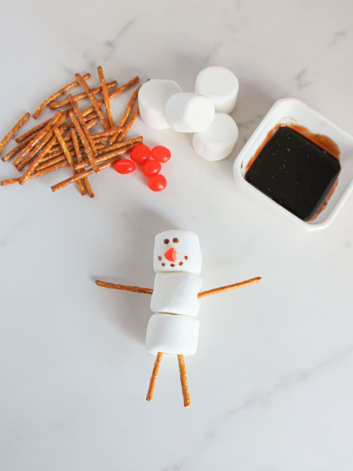 Three marshmallows stacked with pretzel sticks for arms and legs, red candy for buttons, and chocolate sauce for decoration, arranged to resemble a snowman.