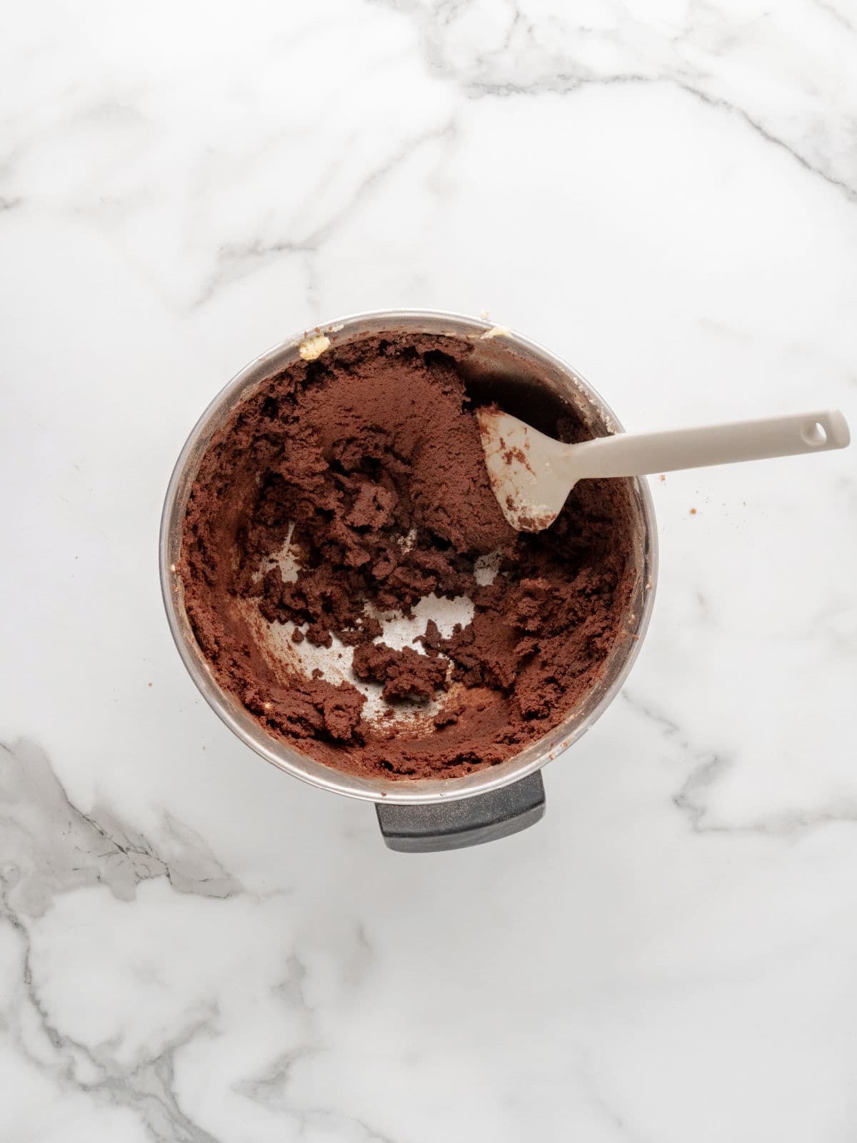A mixing bowl with chocolate cookie dough and a spatula on a white marble surface.