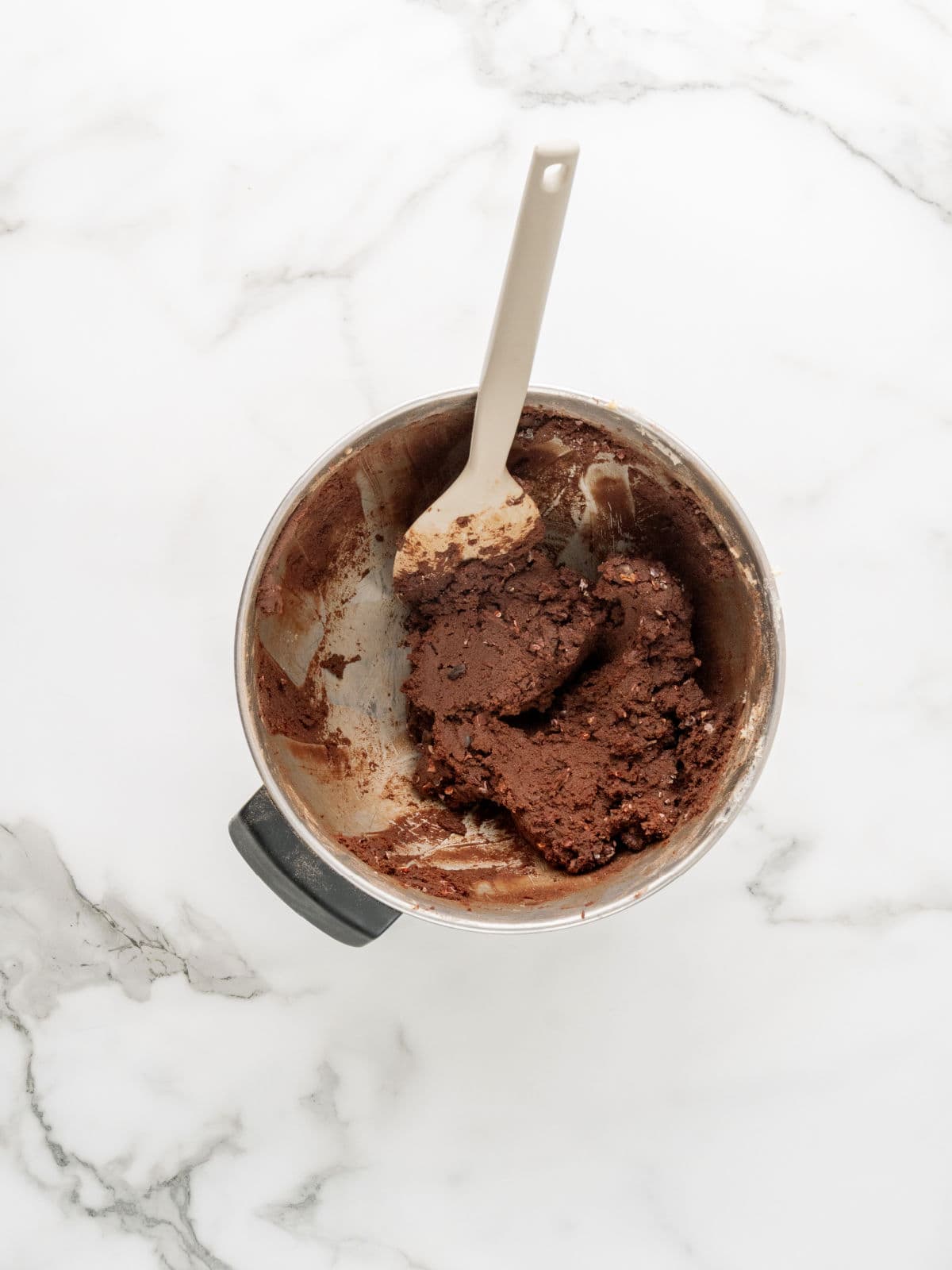 A metal mixing bowl with chocolate cookie dough and a white spatula on a white marble surface.