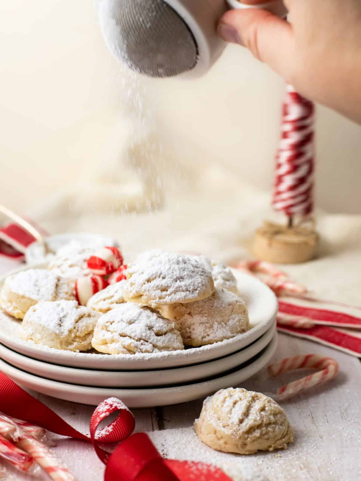 A hand sprinkles powdered sugar over a plate of Christmas cookies, surrounded by candy canes and red ribbon on a white surface.
