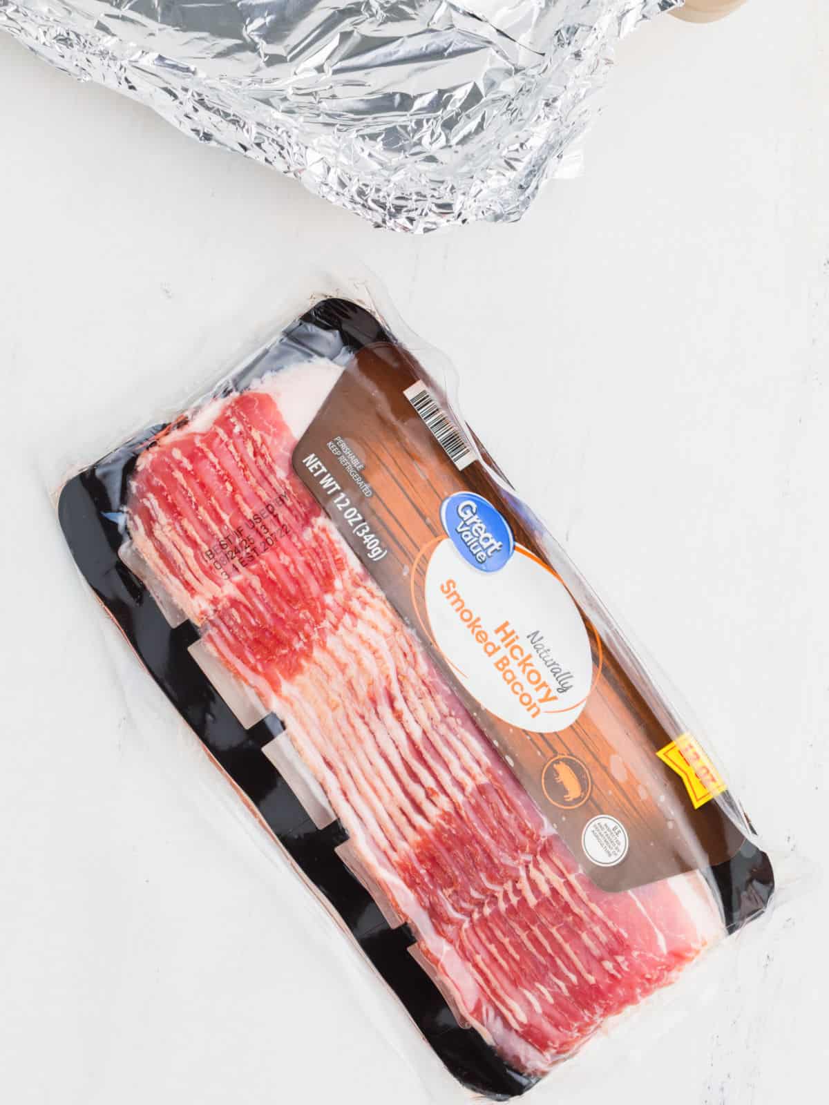 A sealed package of Great Value Hickory Smoked Bacon rests on a white surface next to a foil-covered tray.