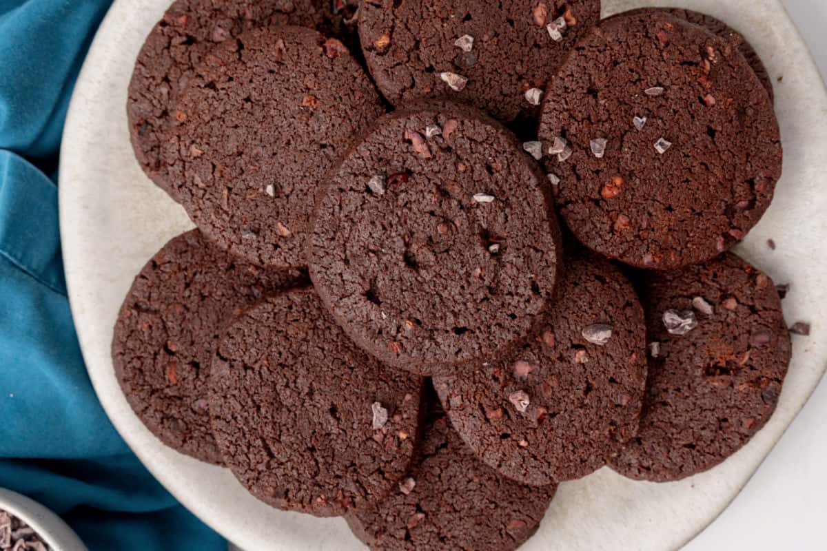 A group of chocolate cookies is arranged on a surface near a white cup of coffee and a bowl of cocoa nibs. One cookie is partially bitten.