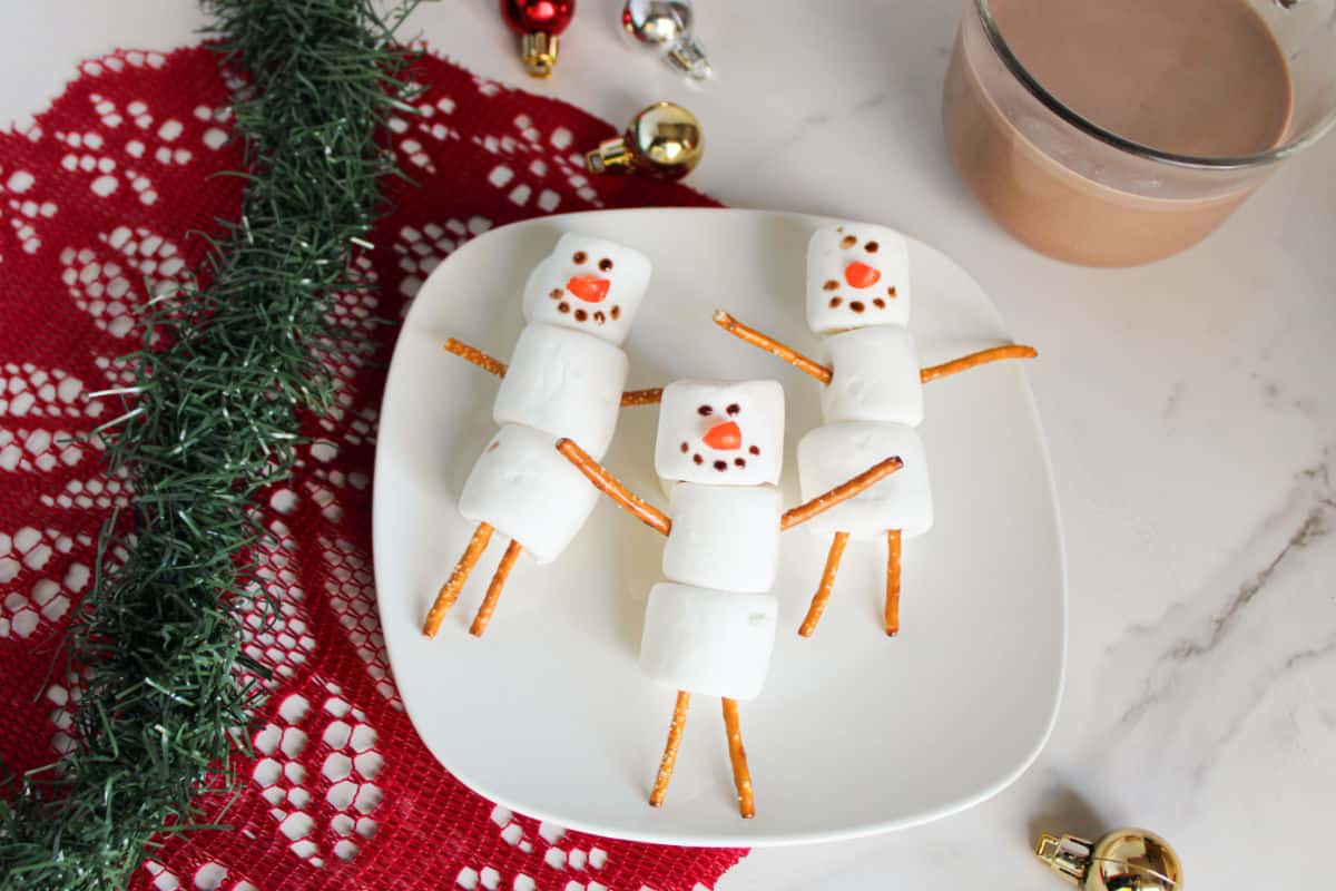 Three marshmallow snowmen with pretzel arms and legs, decorated with chocolate and candy, on a white plate next to holiday decorations and a glass of hot chocolate.