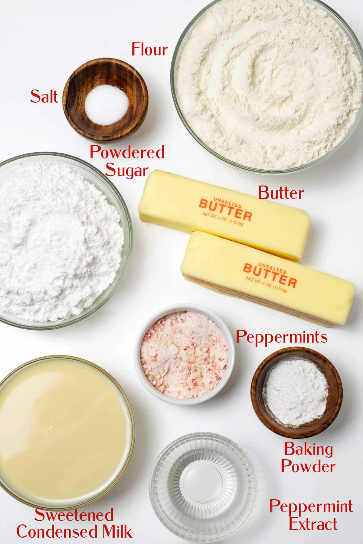 Ingredients for baking peppermint cookies are arranged on a white surface, including flour, butter, powdered sugar, salt, crushed peppermints, baking powder, sweetened condensed milk, and peppermint extract.