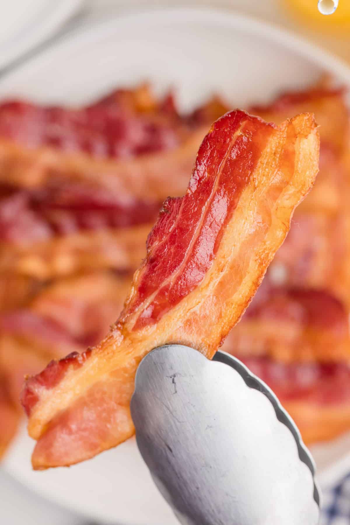 A piece of cooked bacon is held by metal tongs, with more bacon slices on a plate in the blurred background.