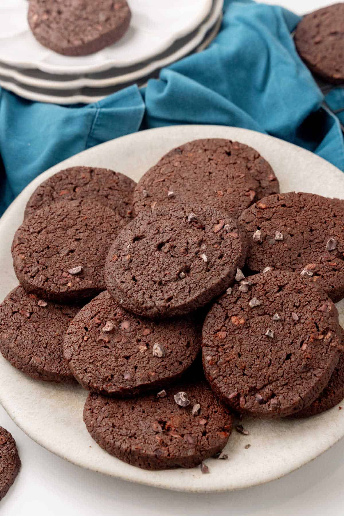 A plate of round, dark chocolate cookies is arranged on a white surface, with a blue cloth and stacked plates in the background.
