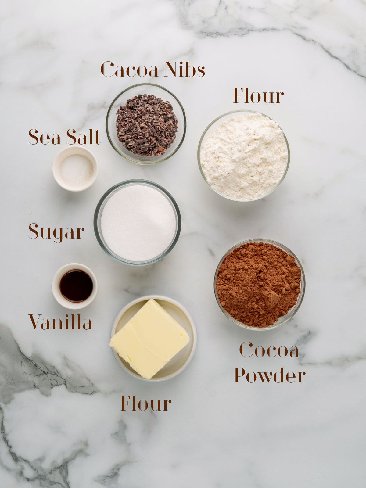 A marble surface with bowls containing cocoa nibs, flour, cocoa powder, sugar, sea salt, vanilla extract, and butter, each labeled with text.