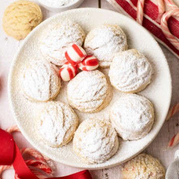 A plate of snowball cookies dusted with powdered sugar, with three red and white striped candies in the center, surrounded by festive holiday ribbons and candy canes.