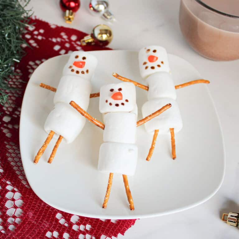 Three snowmen made from marshmallows and pretzel sticks are arranged on a white plate, with holiday decorations and a mug of hot chocolate in the background.