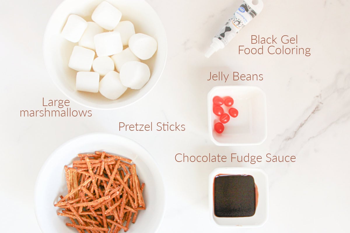 A top-down view of large marshmallows, pretzel sticks, chocolate fudge sauce, red jelly beans, and black gel food coloring in separate bowls on a white surface.