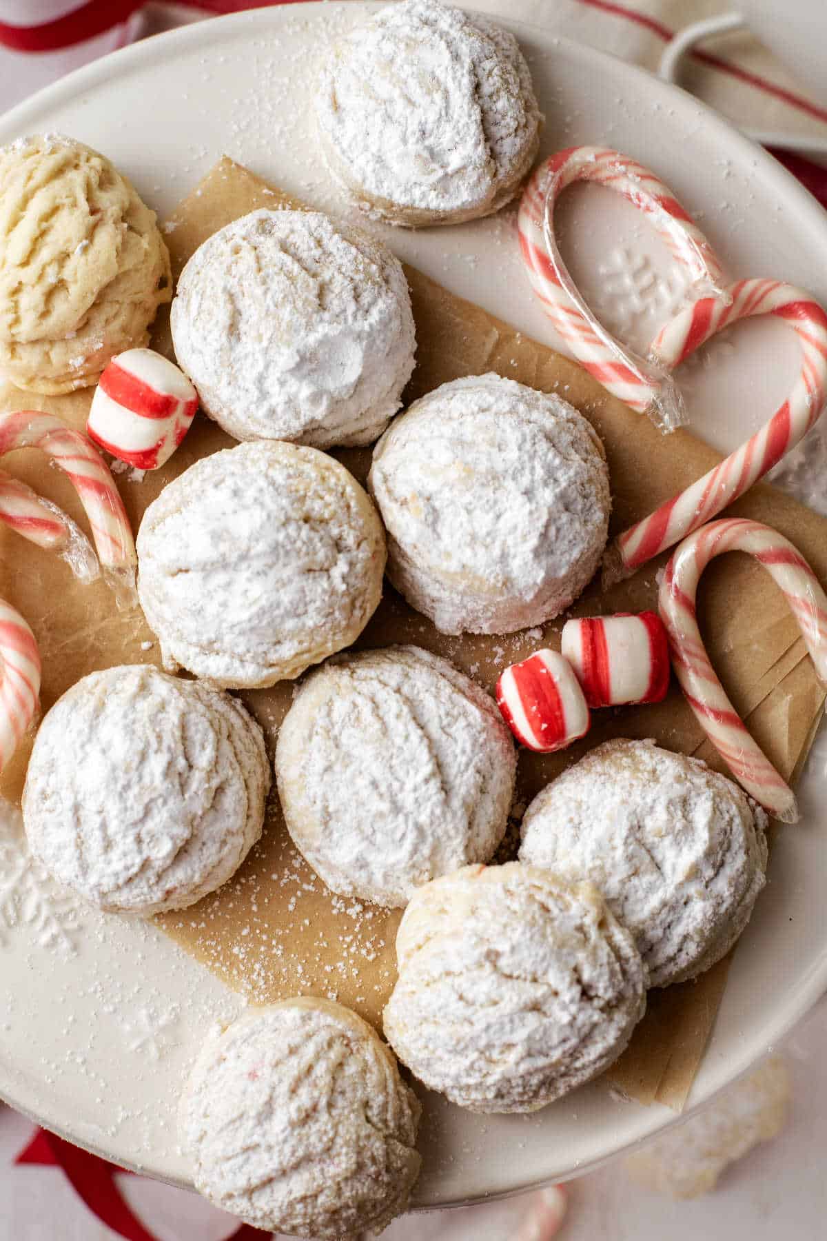 A plate with round powdered sugar cookies, peppermint candies, and candy canes arranged on parchment paper.