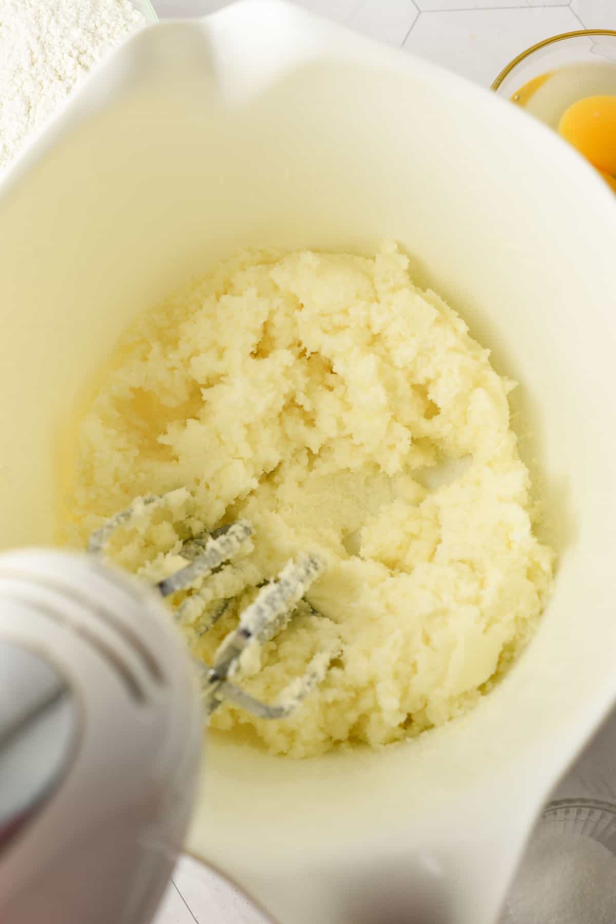 Electric mixer creaming butter and sugar in a white mixing bowl, with eggs and flour visible nearby on a countertop.