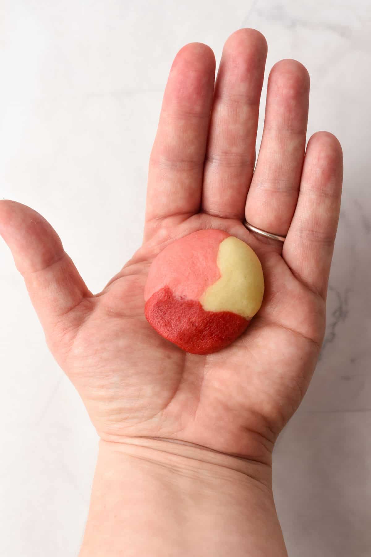 A hand holding a round, tri-colored cookie dough with red, pink, and yellow sections against a light background.