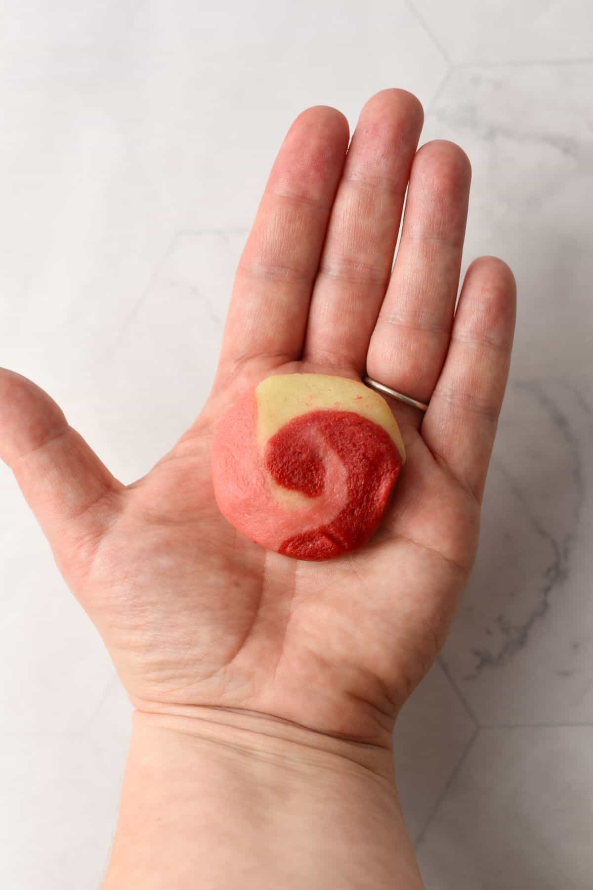 A left hand holds a small round cookie with a red and beige swirl pattern against a light, textured background.