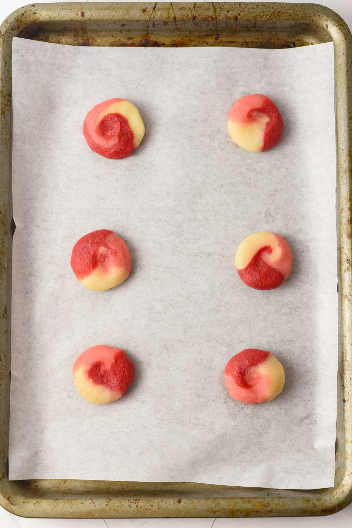 A baking sheet lined with parchment paper holds six unbaked red and white swirled cookies arranged in two columns.