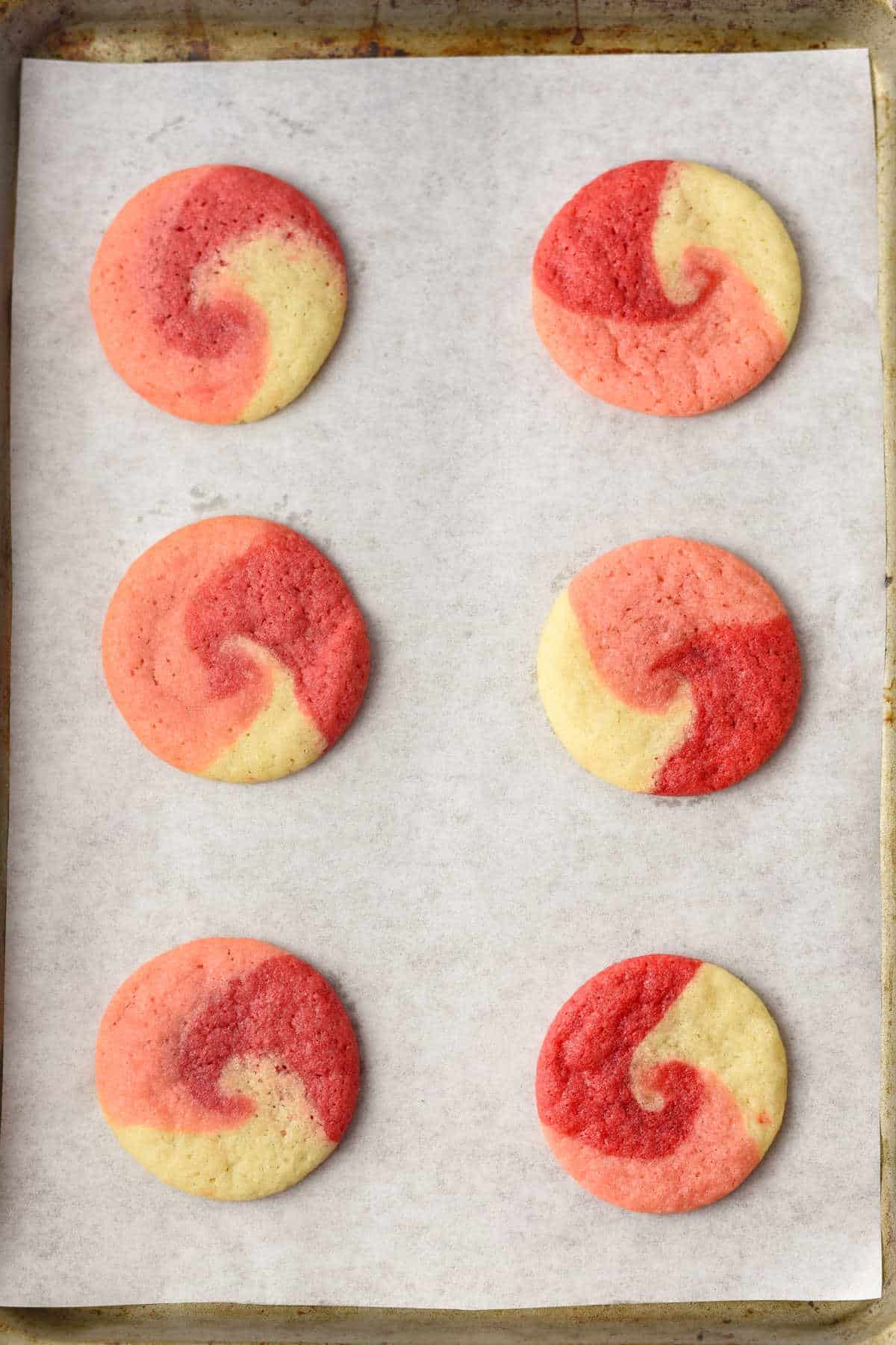 Six red and white swirled sugar cookies are arranged in two columns on a parchment-lined baking sheet.