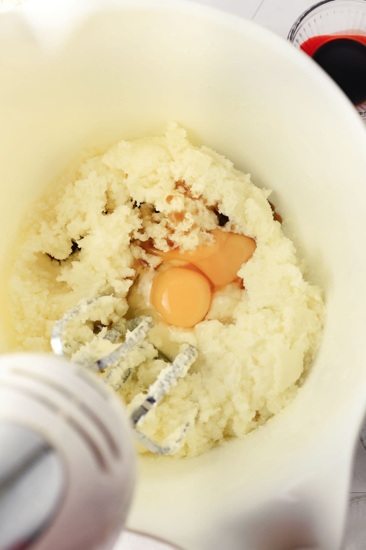 A hand mixer in a bowl with creamed butter and sugar, two raw eggs, and vanilla extract being prepared for baking.