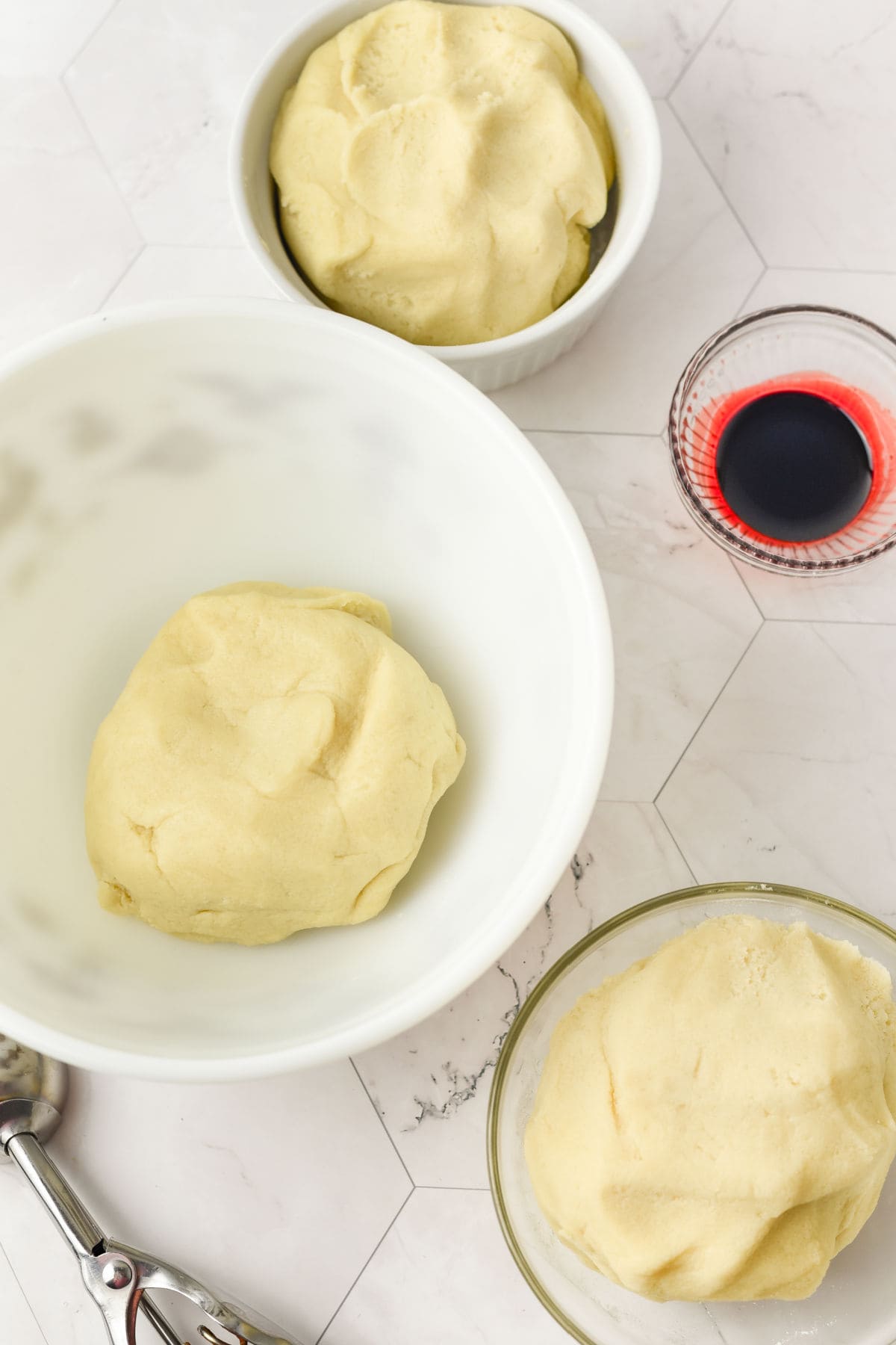 Three bowls of dough and a small glass of red liquid are arranged on a white hexagonal-tiled surface with a metal scoop nearby.
