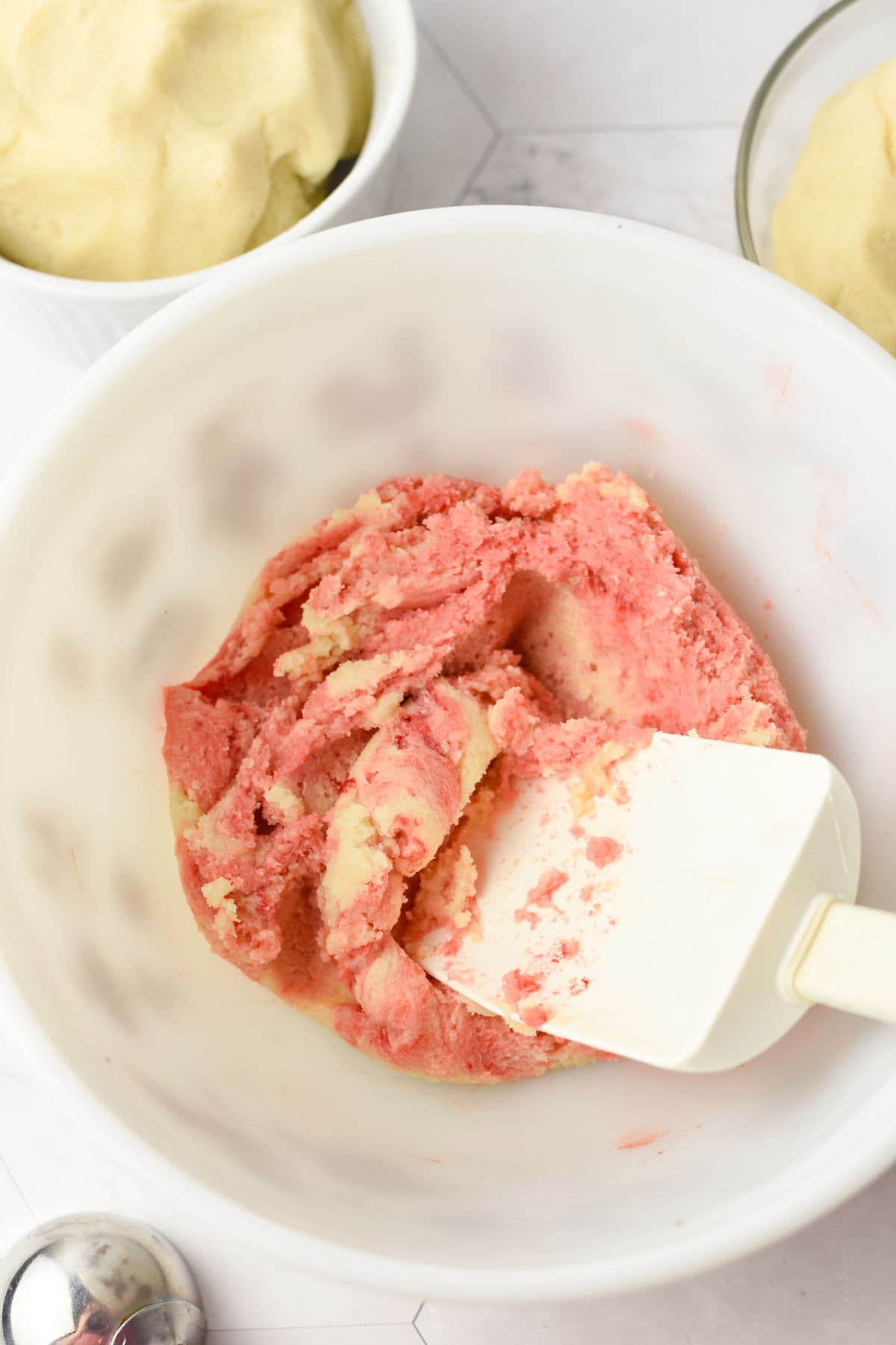 A white mixing bowl with pink and white dough being mixed by a white spatula, with two small bowls of dough in the background.
