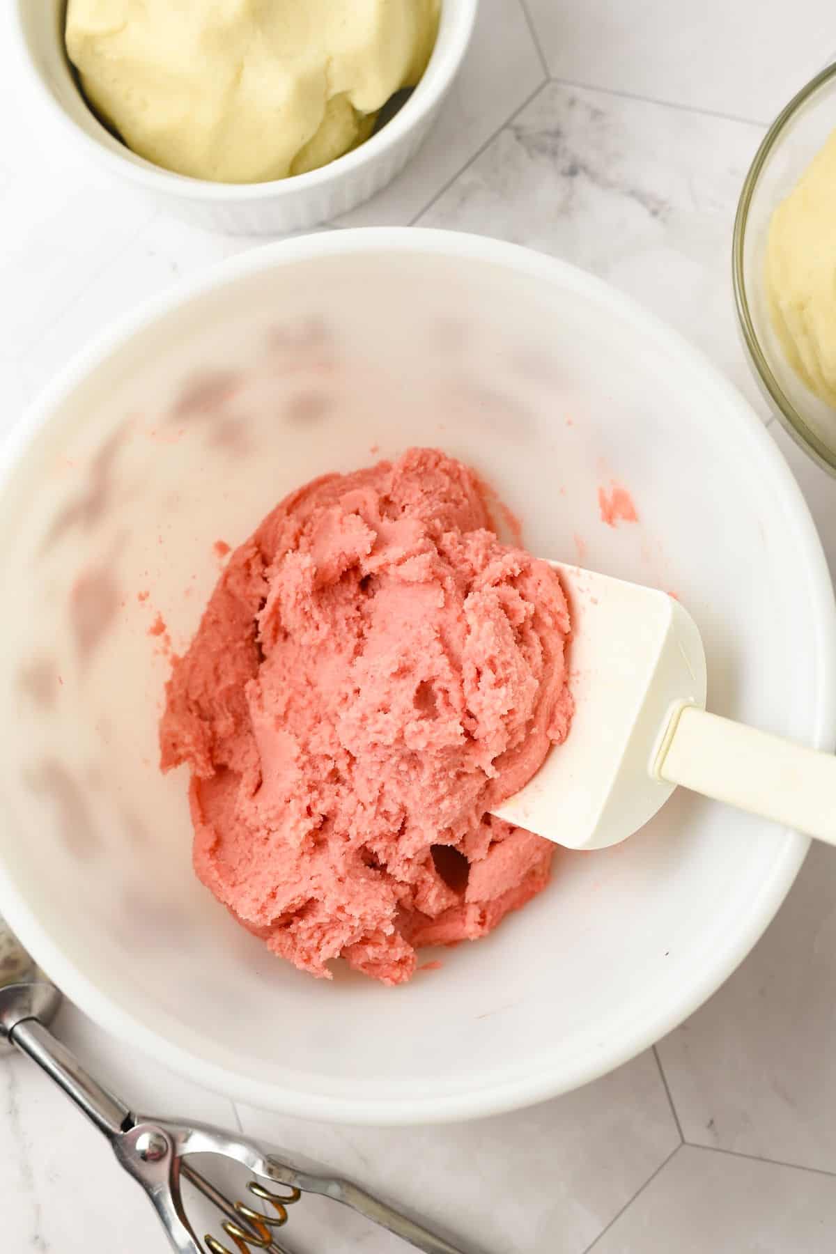 A white mixing bowl contains pink sugar cookie dough being stirred with a white spatula. Nearby are bowls of uncolored dough and a metal cookie scoop on a white surface.