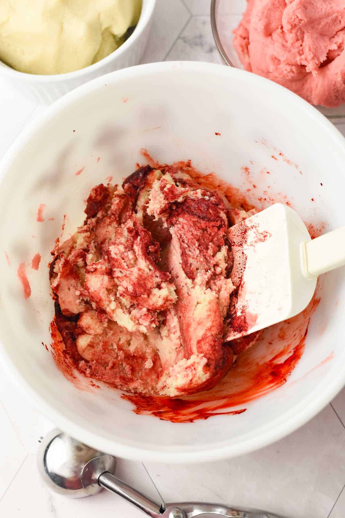 A white bowl with a spatula mixing vanilla and strawberry ice cream, with an ice cream scoop and bowls of plain ice creams nearby on a white surface.