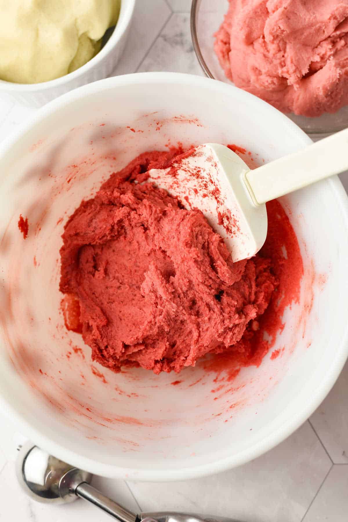 A white mixing bowl containing red cookie dough and a white spatula, with additional bowls of dough in the background on a white surface.