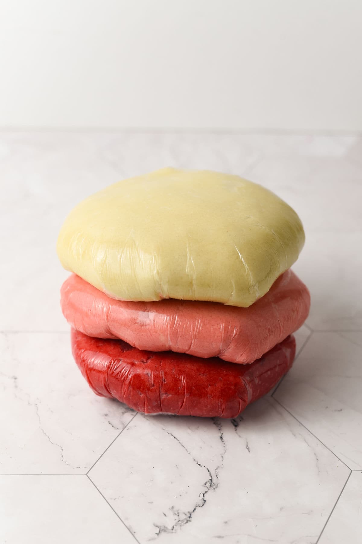 Three discs of dough wrapped in plastic wrap, stacked on a light-colored tiled surface; the dough is yellow, pink, and red from top to bottom.